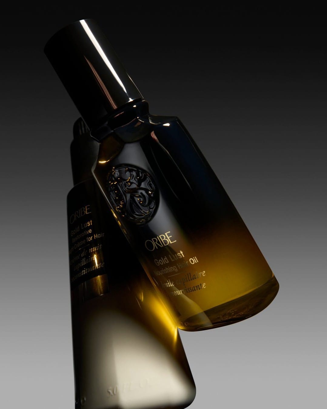 Oribe Gold Lust duo product photo: black glass, amber gold oil and ivory cream, glass, dramatic light